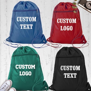 May include: Four drawstring backpacks in blue, red, green, and black. Each bag has a zippered pocket and white text that reads "CUSTOM TEXT" or "CUSTOM LOGO". The bags are on a white wood-grain surface.