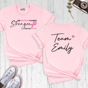 May include: Two pink t-shirts with text. One shirt says "The Comeback is Always Stronger Than The Setback" with a pink ribbon. The other shirt says "Team Emily" with a pink heart. White sneakers are visible.