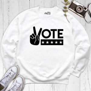 May include: White sweatshirt with a black peace sign and the word VOTE in all caps. Five black stars are below the word VOTE.