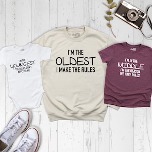 May include: Three shirts with different sayings about being the youngest, oldest, and middle child. The youngest shirt is white, the oldest shirt is beige, and the middle shirt is maroon.