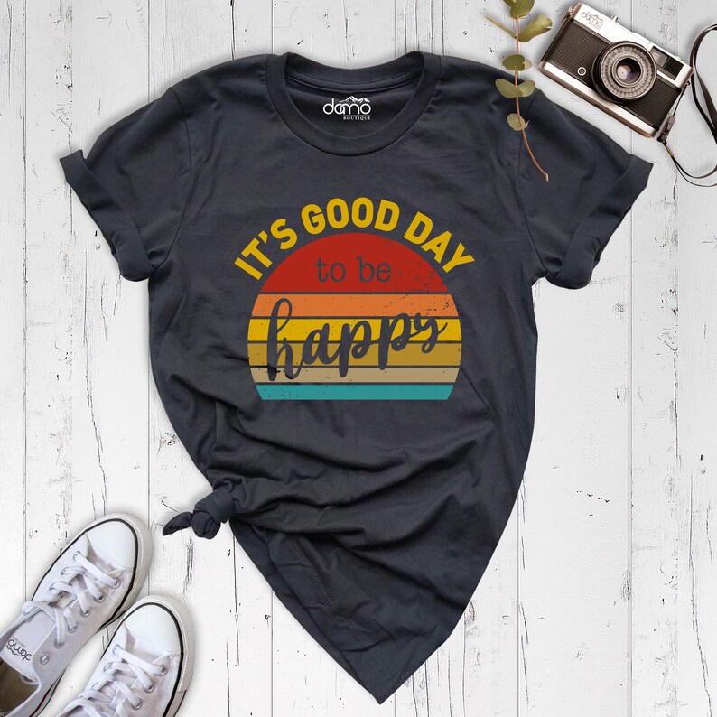 May include: A dark gray t-shirt with a vintage-style graphic that says "It's good day to be happy". The graphic features a sunset with a rainbow of colors.