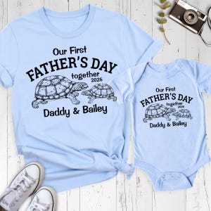 May include: Light blue t-shirt and onesie set. The shirt and onesie feature the text "Our First FATHER'S DAY together 2026 Daddy & Bailey" with a turtle graphic. The set is designed for Father's Day.