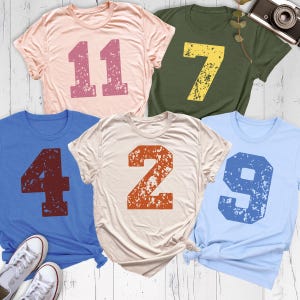May include: Five short-sleeved t-shirts in various colors: pink, olive green, blue, cream, and light blue. Each shirt features a large, distressed number in a contrasting color: 11, 7, 4, 2, and 9. The shirts are casual and likely made of soft fabric.