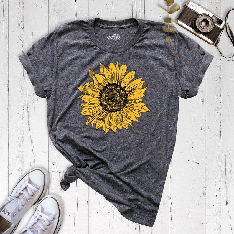 以下が含まれることがあります： 黄色のひまわり柄のグレーのTシャツ。ひまわりはヴィンテージスタイルで描かれています。