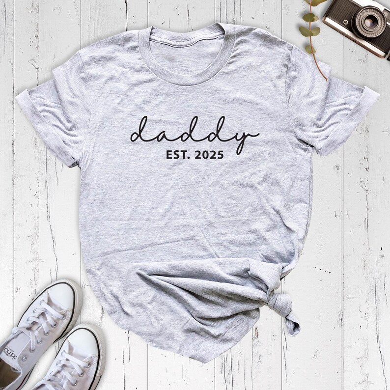 May include: Gray t-shirt with the text "daddy est. 2025" in black script font.