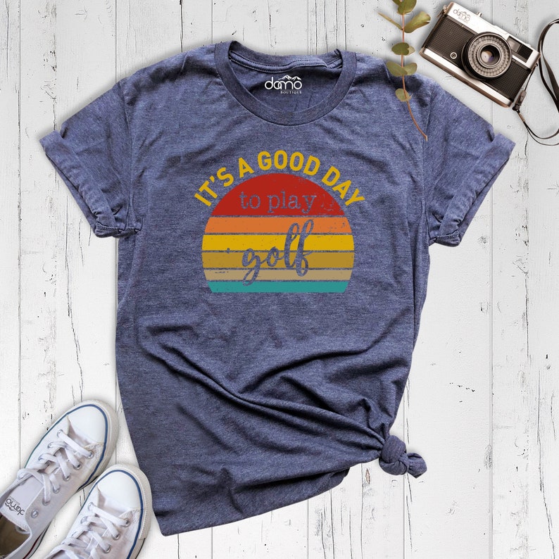 May include: A navy blue t-shirt with a vintage-style sunset graphic and the text "It's a good day to play golf".