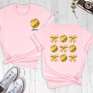 May include: Two pink t-shirts with a softball design. The front shirt has a single softball with a bow and the text "Jessica". The back shirt has a pattern of six softballs with bows.