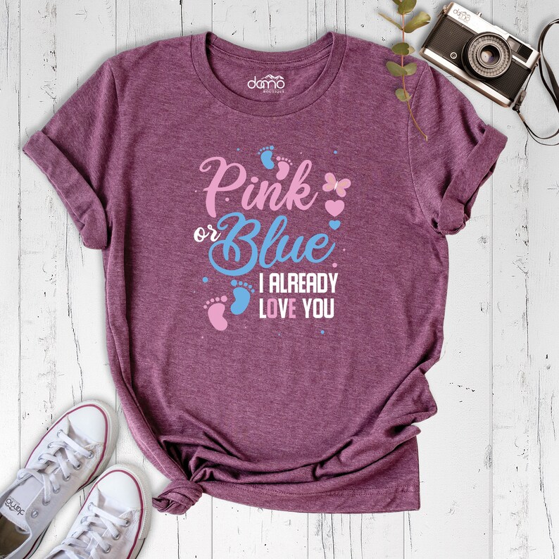May include: A heather purple t-shirt with the text "Pink or Blue I Already Love You" in blue and pink lettering. The shirt features a pink butterfly and pink and blue footprints.
