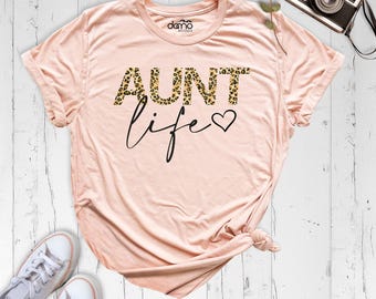 May include: A light pink short-sleeve t-shirt featuring the text AUNT in a leopard print pattern and the word life in black cursive script followed by a small black heart symbol. The shirt has a crew neckline and a knotted hem detail.