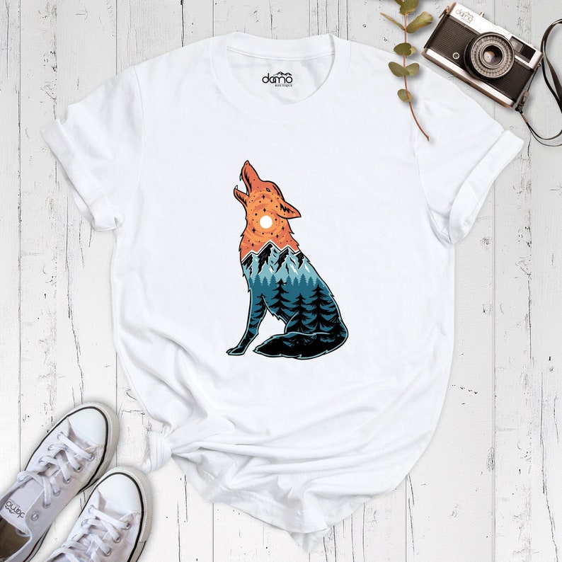 Puede incluir: Camiseta blanca con un gr&aacute;fico de un lobo aullando con un paisaje de monta&ntilde;a dentro de su silueta. El lobo est&aacute; mirando hacia la izquierda y las monta&ntilde;as est&aacute;n en tonos de azul, verde y naranja.