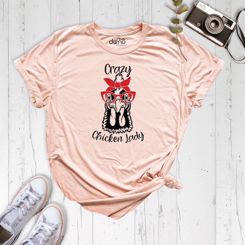K&ouml;nnte beinhalten: Ein pfirsichfarbenes T-Shirt mit einer Schwarzwei&szlig;illustration eines Huhns, das eine rote Brille und ein rotes Bandana tr&auml;gt. Der Text "Crazy Chicken Lady" ist unter der Illustration aufgedruckt.