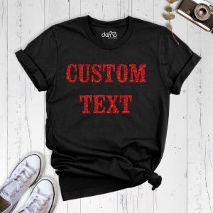 以下が含まれることがあります： 中央に赤いグリッターのテキスト「CUSTOM TEXT」がプリントされた黒の半袖Tシャツ。クルーネックで、襟の内側にブランドロゴがプリントされています。白いスニーカーと一緒に平置きされた状態で撮影されています。