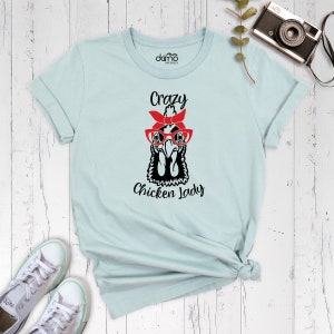 K&ouml;nnte beinhalten: Hellblaues T-Shirt mit einer Schwarz-Wei&szlig;-Illustration eines Huhns, das eine rote Brille und ein rotes Bandana tr&auml;gt. Der Text "Crazy Chicken Lady" ist unter der Illustration aufgedruckt.