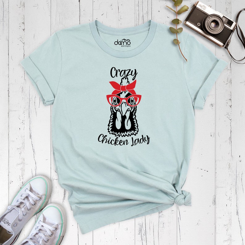 K&ouml;nnte beinhalten: Hellblaues T-Shirt mit einer Schwarz-Wei&szlig;-Illustration eines Huhns, das eine rote Brille und ein rotes Bandana tr&auml;gt. Der Text "Crazy Chicken Lady" ist unter der Illustration aufgedruckt.