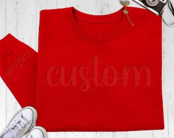 May include: A bright red crewneck sweatshirt featuring the word custom embossed in a tonal red script font across the chest. The sweatshirt has long sleeves with the word custom visible on the cuff and is paired with white sneakers.