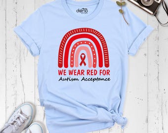 May include: A light blue t-shirt featuring a graphic of a red and pink rainbow with a red awareness ribbon in the center. The text reads WE WEAR RED FOR Autism Acceptance in red and black lettering. The shirt has a crew neck and short sleeves.