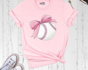 May include: A light pink short-sleeve t-shirt featuring a graphic of a white baseball with pink stitching and a large pink bow on top. The shirt has a crew neckline and the text "domo" printed in small black letters near the collar.