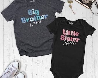 May include: A charcoal gray short-sleeve t-shirt with the text Big Brother David in blue and a black short-sleeve baby bodysuit with the text Little Sister Katie in pink. A pair of white sneakers is positioned at the bottom left corner.