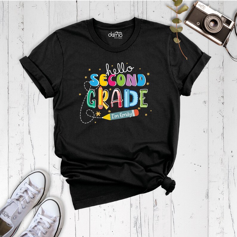 May include: Black t-shirt with the text "hello second grade" in colorful letters and a pencil with the text "I'm Emily" on it.