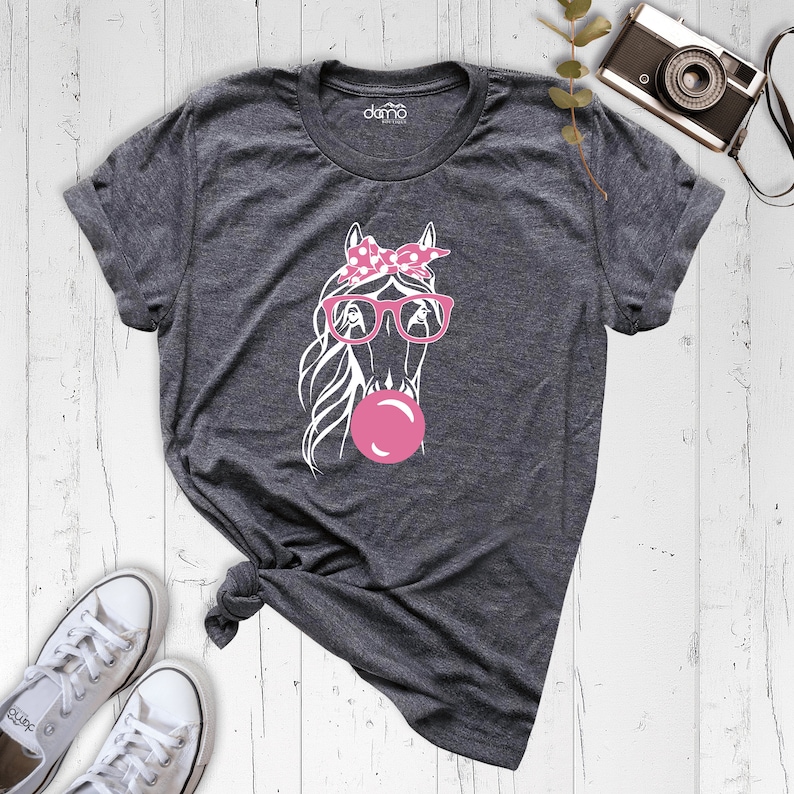 Puede incluir: Una camiseta gris con un gr&aacute;fico blanco de un caballo con gafas rosas y un pa&ntilde;uelo rosa con lunares. El caballo est&aacute; soplando una burbuja rosa.