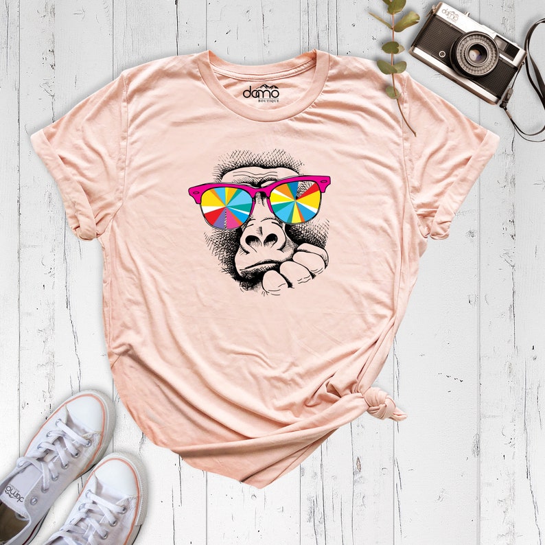 May include: A peach-colored t-shirt with a black and white illustration of a gorilla wearing pink and rainbow sunglasses. The text "domo" is printed on the shirt.