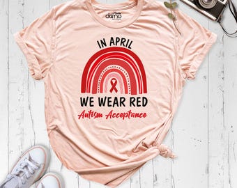 May include: A light peach-colored t-shirt featuring a red rainbow graphic with a central ribbon icon. The text reads: IN APRIL WE WEAR RED Autism Acceptance. The shirt has short sleeves, a crew neckline, and a small logo on the inner collar.