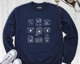 May include: A navy blue crewneck sweatshirt featuring a white line art graphic of nine different vintage cameras arranged in a grid. The interior neck label displays the text damo boutique in white lettering.