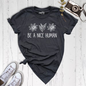 Op de afbeelding: Een donkergrijs T-shirt met witte tekst die "BE A NICE HUMAN" luidt en een afbeelding van drie gestileerde bloemen en bladeren.