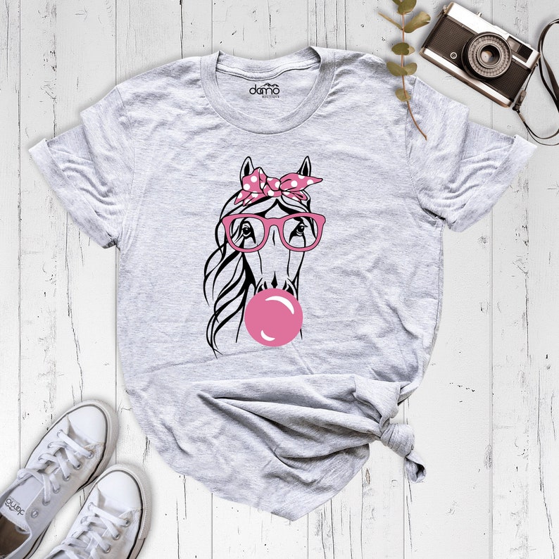 Puede incluir: Una camiseta gris con una ilustraci&oacute;n en blanco y negro de un caballo que lleva gafas rosas y un pa&ntilde;uelo de lunares rosas. El caballo est&aacute; soplando una burbuja rosa.