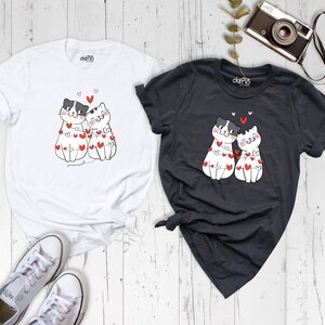 May include: Two white t-shirts with a black and white cartoon image of two cats wearing a string of red hearts. One shirt is white and the other is dark grey.