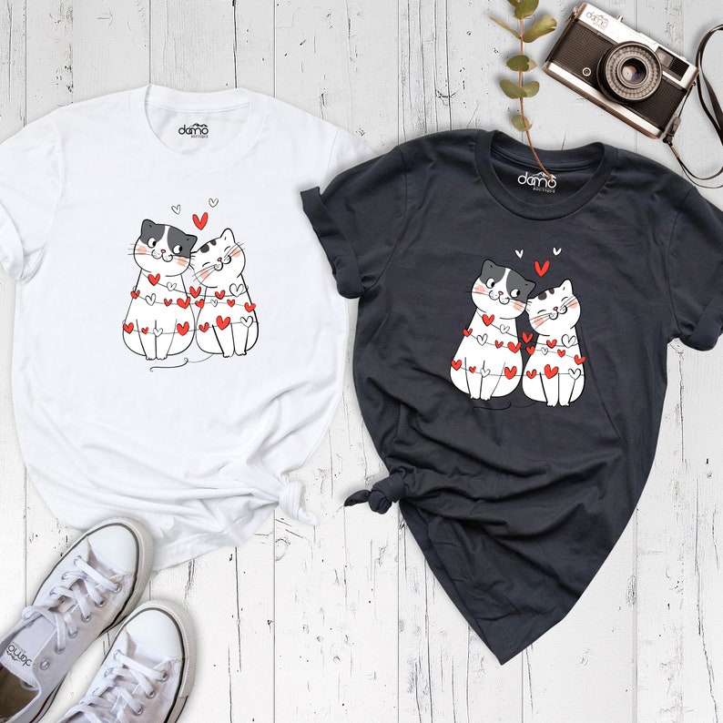 May include: Two white t-shirts with a black and white cartoon image of two cats wearing a string of red hearts. One shirt is white and the other is dark grey.