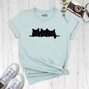 May include: A light blue t-shirt with a black silhouette of three cats peeking over a ledge. The cats are facing forward with their eyes open.