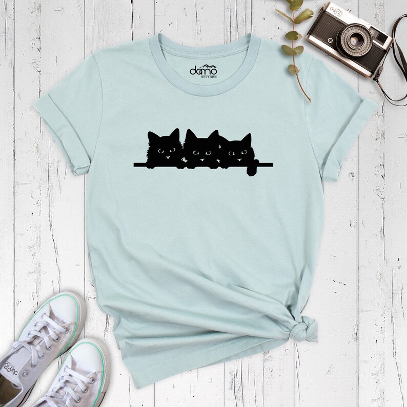 May include: A light blue t-shirt with a black silhouette of three cats peeking over a ledge. The cats are facing forward with their eyes open.
