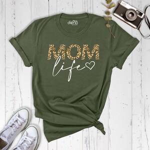 Może przedstawiać: Zielona koszulka z napisem "MOM life" w białym kolorze. Słowo "MOM" jest w wzorze lamparta z małym sercem obok słowa "life".