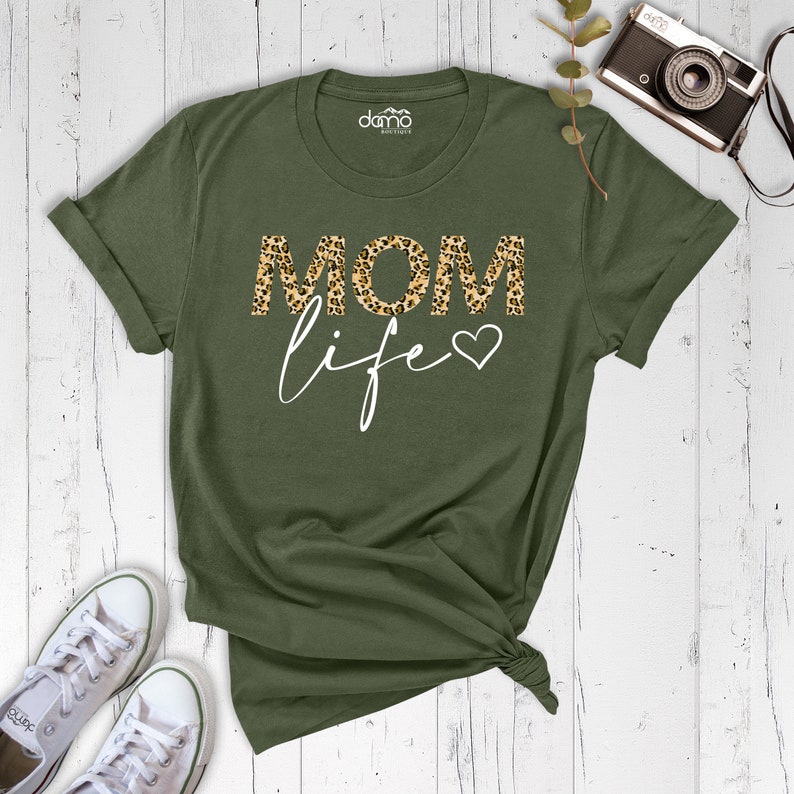 Może przedstawiać: Zielona koszulka z napisem "MOM life" w białym kolorze. Słowo "MOM" jest w wzorze lamparta z małym sercem obok słowa "life".