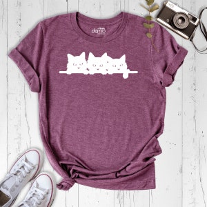 May include: A maroon t-shirt with a white graphic of three kittens peeking over a ledge. The kittens are all facing forward with their ears perked up.