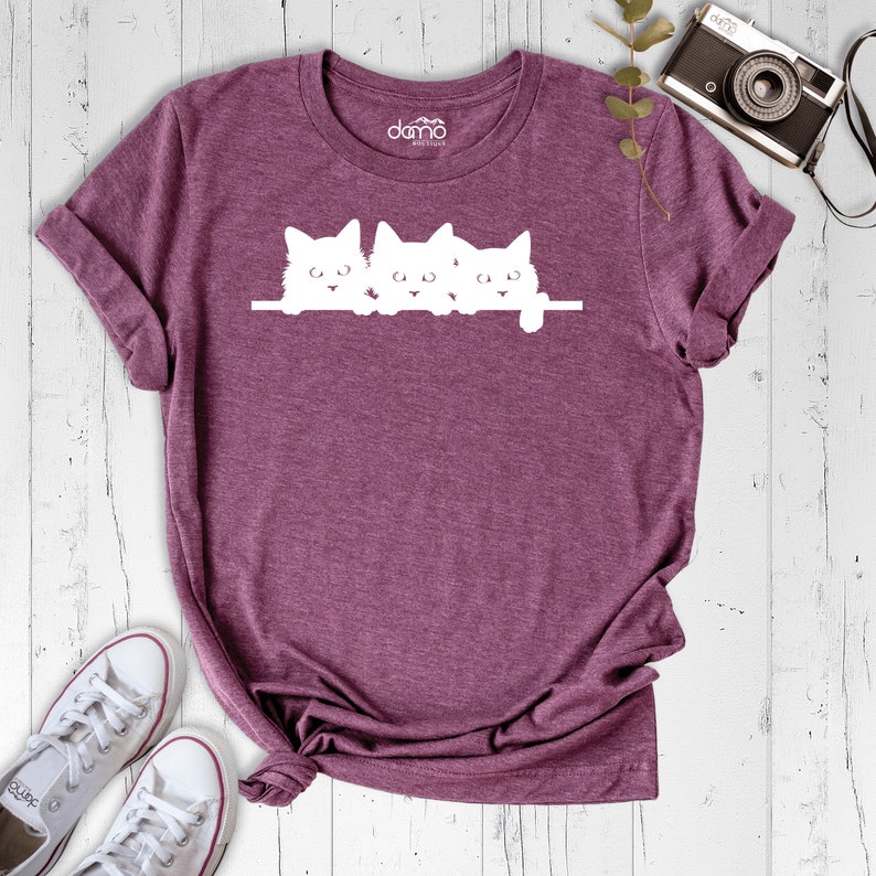 May include: A maroon t-shirt with a white graphic of three kittens peeking over a ledge. The kittens are all facing forward with their ears perked up.