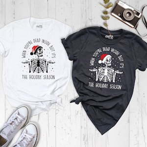 May include: A white t-shirt and a dark gray t-shirt, each featuring a graphic of a skeleton wearing a red Santa hat with the text "WHEN YOU'RE DEAD INSIDE BUT IT'S THE HOLIDAY SEASON" printed in black or white ink on the front of the garments.