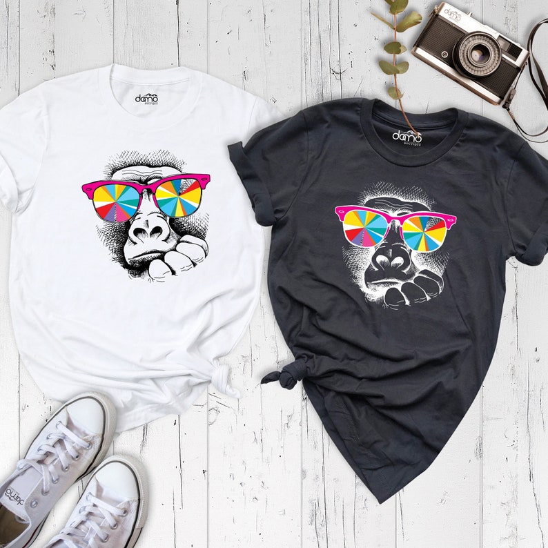 May include: Two t-shirts with a graphic of a gorilla wearing colorful sunglasses. The t-shirts are white and black.