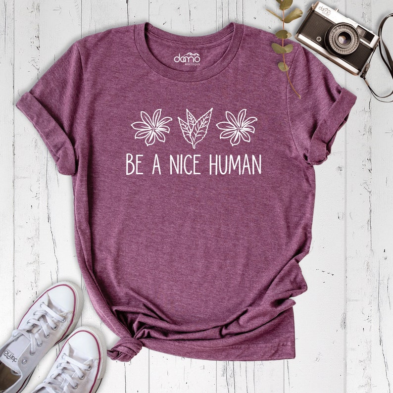Op de afbeelding: Een maronkleurig T-shirt met een wit grafisch ontwerp van drie gestileerde bloemen en bladeren. De tekst "BE A NICE HUMAN" is in wit onder de bloemen gedrukt.