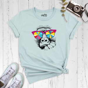 May include: A light blue t-shirt with a graphic of a gorilla wearing colorful sunglasses. The gorilla is looking to the side and has a thoughtful expression.
