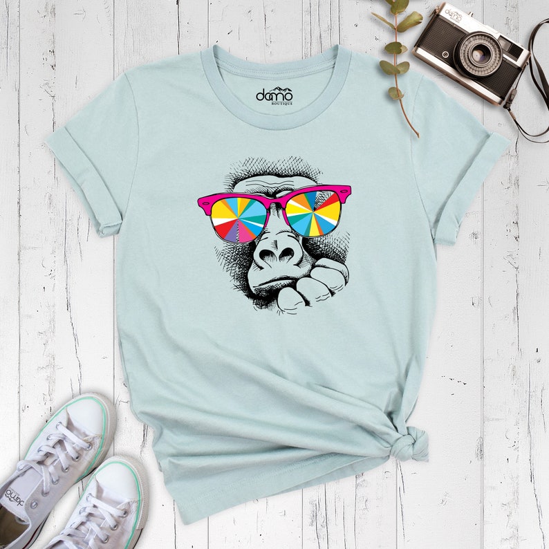 May include: A light blue t-shirt with a graphic of a gorilla wearing colorful sunglasses. The gorilla is looking to the side and has a thoughtful expression.