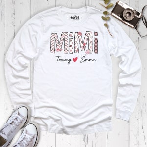 May include: A white long-sleeved shirt with the word "MIMI" in a decorative font, featuring pink and black patterns. Below, the names "Tommy" and "Emma" are written with a heart symbol. White trainers and a vintage camera are also visible.
