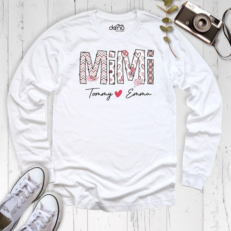 May include: A white long-sleeved shirt with the word "MIMI" in a decorative font, featuring pink and black patterns. Below, the names "Tommy" and "Emma" are written with a heart symbol. White trainers and a vintage camera are also visible.