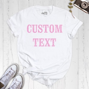 May include: A white short-sleeved t-shirt featuring the words CUSTOM TEXT printed in a pink glittery font across the chest. The shirt has a crew neckline and a brand logo printed on the inside collar area. The fabric is plain white cotton.