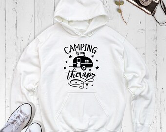 May include: A white hooded sweatshirt featuring a black graphic print on the chest. The design includes a camper trailer icon surrounded by stars and hearts, with the text "CAMPING is my therapy" written in a mix of bold and script fonts.