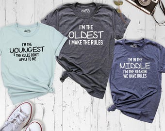 May include: Three short-sleeve t-shirts in light blue, dark gray, and navy blue. The light blue shirt reads I'M THE YOUNGEST THE RULES DON'T APPLY TO ME. The dark gray shirt reads I'M THE OLDEST I MAKE THE RULES. The navy blue shirt reads I'M IN THE MIDDLE I'M THE REASON WE HAVE RULES.