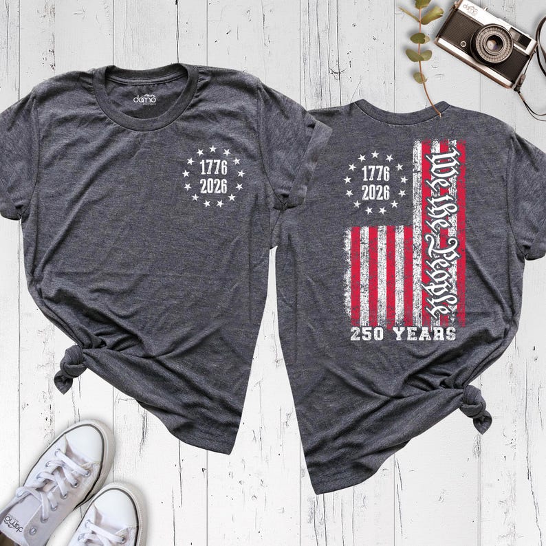 May include: A charcoal grey heather t-shirt featuring a front chest print of a circle of stars with the text 1776 2026. The back print displays a distressed American flag graphic with the text We the People and 250 YEARS below the flag.