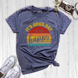May include: A navy blue t-shirt with a vintage-style graphic that says "It's good day to be happy" with a sunset design.