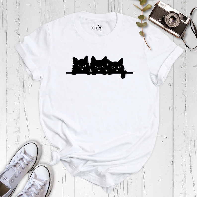 May include: White t-shirt with a black silhouette of three cats peeking over a surface. The cats are in a row, with their heads and paws visible.
